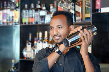 Professional african bartender male holding in hands a shaker with a fresh delicious cocktail. Bartender shaking a cocktail shaker as she stands behind the bar mixing a drink for a client. Close-up