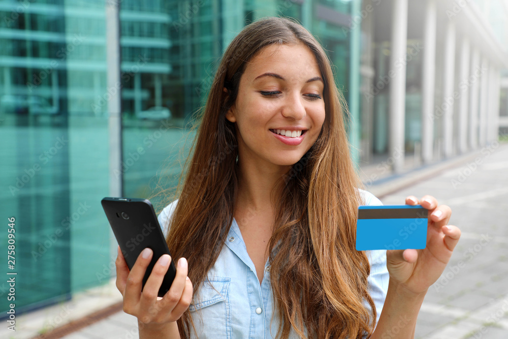 Wall mural Portrait of a happy woman buying online with a smart phone and showing her credit card outdoors