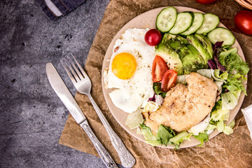 Served healthy breakfast with fried eggs and vegetable Top view of plate of tasty fried eggs with chicken and avocado with colorful vegetable salad placed next to knife and fork on kitchen table