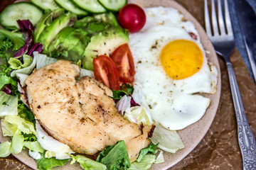 Served healthy breakfast with fried eggs and vegetable Top view of plate of tasty fried eggs with chicken and avocado with colorful vegetable salad placed next to knife and fork on kitchen table