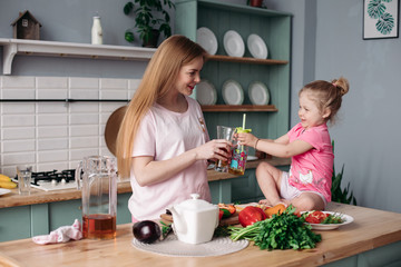 Beautiful young mother cooking dinner on kitchen with her little cute kid. Daughter sitting on table and drinking juice together with mom. Happy woman making lunch from vegetables together with child.