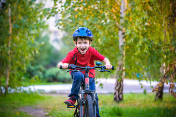 A small white Toddler boy in a protective helmet on his head sits on a children's bicycle. Toddler on a two-wheeled red bicycle looks forward. A sly smile on the child's face