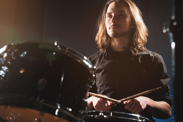 Fototapeta na wymiar Portrait of a long-haired drummer with chopsticks in his hands sitting behind a drum set. Low key. Concepts of the creative freedom of the millenial generation