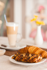 A croissant and a chocolate bun on a white plate in the foreground, a tall glass of latte and a white vase with yellow chrysanthemums in the background in the cafe. Morning in the cafe.