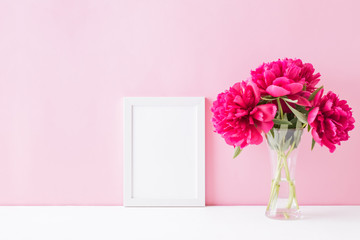 Mockup with a white frame and red peonies in a vase on a pink background