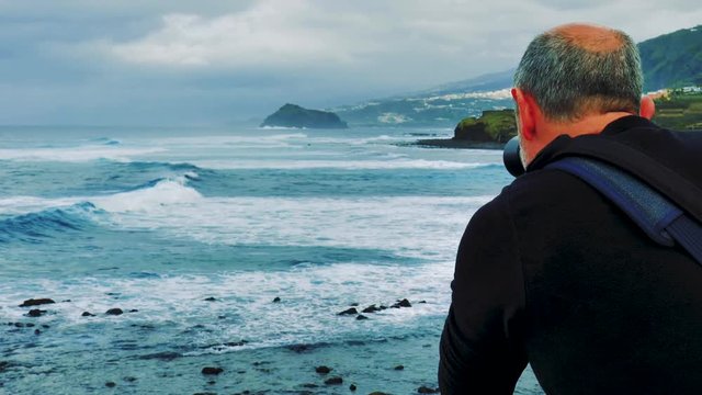 Man taking pictures of the ocean and waves pounding the rocky shoreline of Tenerife. Cloudy day with high surf. Dramatic view of coastal conditions and the Canary Islands landscape.