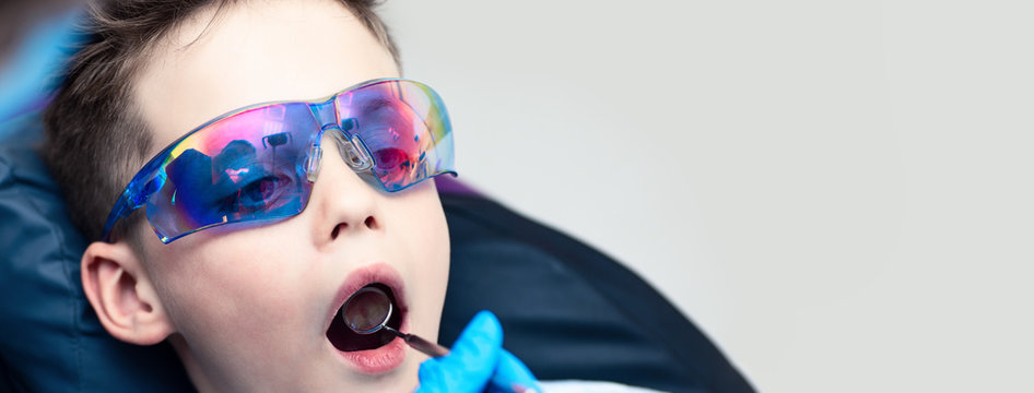 A boy with goggles in the dental chair. The doctor examines the oral cavity with a special dental mirror