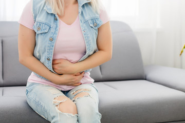 Asian girl having period is sitting on sofa and feeling much of painful on her stomach. It is called women's pain or menstrual syndrome. She holds onto her stomach and curls through spasms.