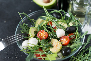 plate with vegetable salad, arugula, tomato, mozzarella, avocado slices, sesame, olives, dark background