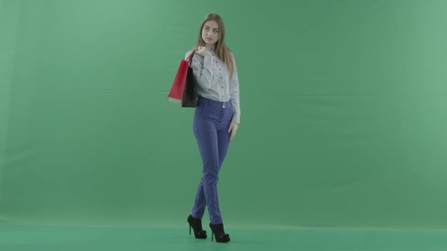 Woman with her purchases is posing for camera. Lady with straight brunette hair is wearing casual shirt and jeans. Female shopper holding shopping bags is standing on green background in a studio.