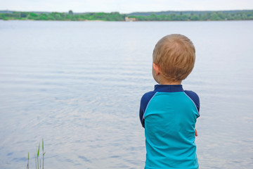 Little boy 3 years old is standing near the water, on the lake shore, dressed in swimming shorts and T-shirt. View from the back. Space for text