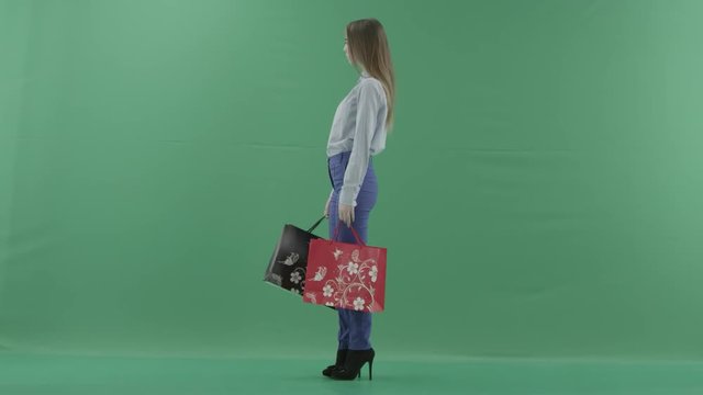 Woman with her purchases is posing for camera. Lady with straight brunette hair is wearing casual shirt and jeans. Female shopper holding shopping bags is standing on green background in a studio.