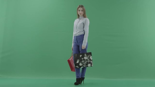 Woman with her purchases is posing for camera. Lady with straight brunette hair is wearing casual shirt and jeans. Female shopper holding shopping bags is standing on green background in a studio.