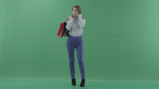 Woman with her purchases is posing for camera. Lady with straight brunette hair is wearing casual shirt and jeans. Female shopper holding shopping bags is standing on green background in a studio.