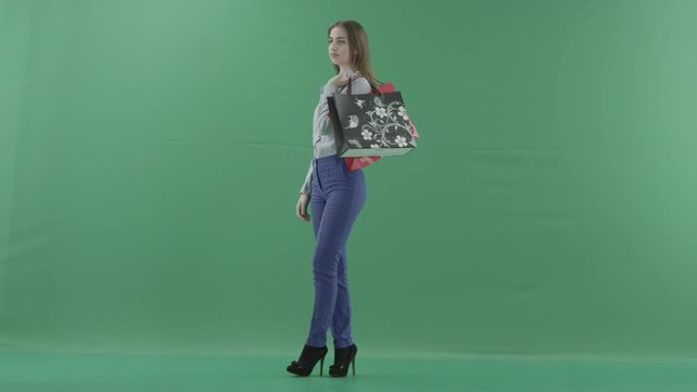 Woman with her purchases is posing for camera. Lady with straight brunette hair is wearing casual shirt and jeans. Female shopper holding shopping bags is standing on green background in a studio.