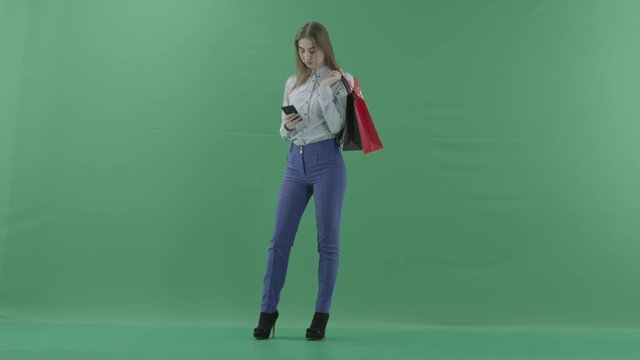 Woman with her purchases is surfing the internet or messaging. Lady with straight brunette hair is wearing casual shirt and jeans. Female shopper holding shopping bags is standing on green background