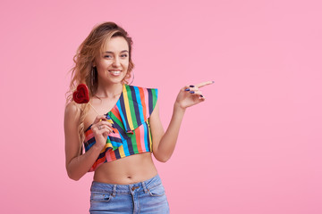 Fashion portrait of pretty sweet young smiling woman having fun with lollipop over pink background. It shows a finger to the right side. Copy space for text