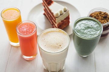 a red velvet cake on the background of glasses with various colored drinks (juice, coffee, smoothie, milkshake) standing on a white wooden table. A concept for a baker's dessert, a cafe, a restaurant