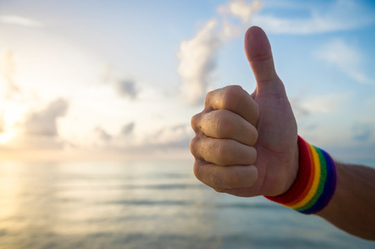 Hand of athlete giving thumbs up with gay pride rainbow colors wristband against soft sky sea horizon
