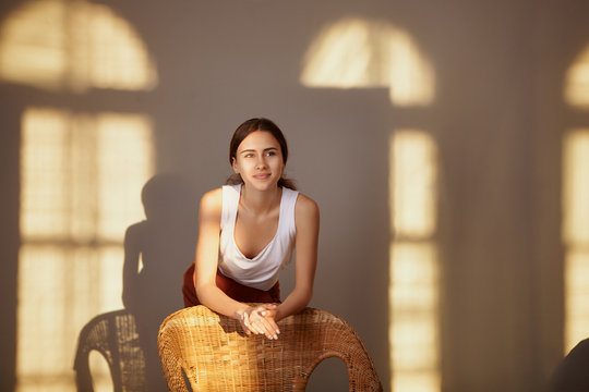 Portrait of cheerful cute young European female posing in spacious living room illuminated with golden hour sunlight placing elbows on back of stylish woven armchair having joyful happy smile
