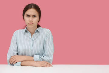 Serious displeased student girl wearing striped shirt keeping elbows on desk, frowning, being angry...