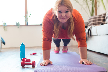 Chubby woman sport at home standing on arms on mat looking camera happy