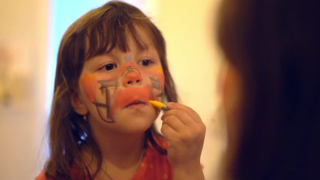 Portrait of little caucasian girl paints on face with face paint in front the mirror