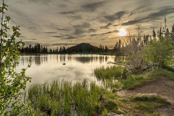 Backlit Elerald Mountain reflected in ripples of Sprague Lake with dramatic clouds and bright sun in the background