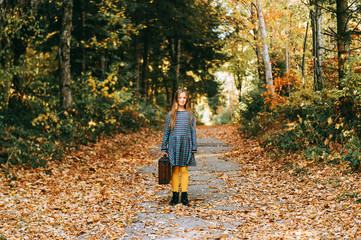 Autumn portrait of pretty young girl with old vintage suitcase, kid model posing outdoor in the forest, wearing plaid dress and yellow tights