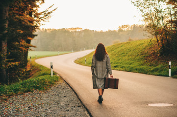 Beautiful woman walking away down the road, wearing long coat, holding old vintage suitcase, back view