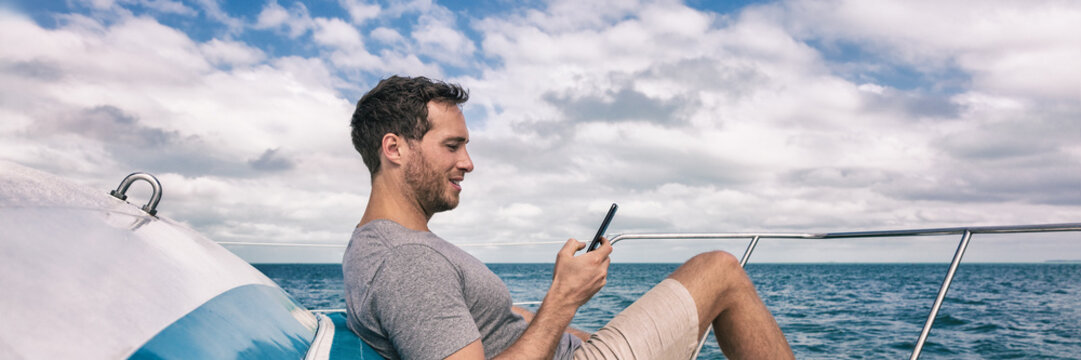 Yacht luxury lifestyle young man using cellphone banner panorama. Person relaxing on deck texting sms message on mobile phone under the sun summer holidays.