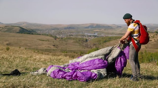 man beard mustache striped white-yellow T-shirt green hat jeans with red backpack collects carries off purple gray parachute against the background mountains, city lake in summer