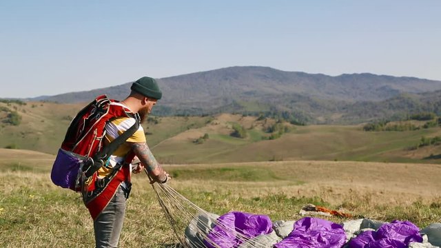 man beard mustache striped white-yellow T-shirt green hat jeans with red backpack collects carries off purple gray parachute against the background mountains, city lake in summer