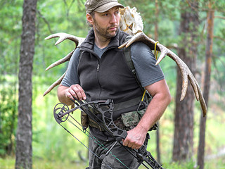 A hunter with a bow in the woods carries moose horns on his back and looking by side