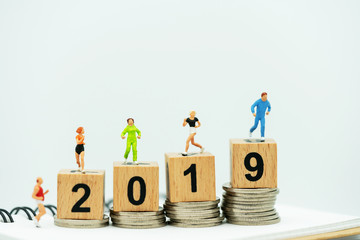 Miniature people running on a wooden box with number 2019 and coins.