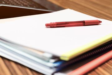 Soft focus in vintage style of red pen on pile of unfinished paperwork stacked in archive with colorful plastic slide binder bars and laptop nearby. Business and education concept.