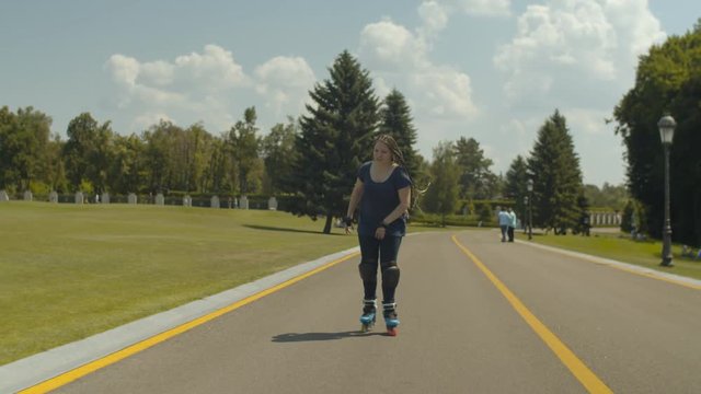 Smiling female with long afro-braids rollerskating forward at speed in summer park lane. Cheerful woman in protective outfit spending outdoor leisure rollerblading along public park in summer.