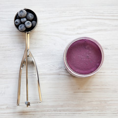 Blueberry ice cream sorbet in a plastic jar, icecream scoop with frozen blueberries on a white wooden background, view from above. Top view.