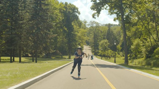Happy fearless female in protective equipment rollerblading at speed up and down park alley enjoying freedom of extreme ride. Joyful woman riding roller blades in green park on sunny windy day.