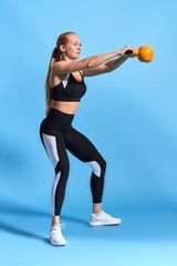 young attractive female sportswoman doing kettle bell exercise on blue background. Fitness girl working out. Crossfit, full length side view photo
