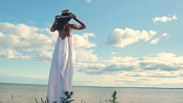 a beautiful slender girl in a white dress takes off her straw hat and spreads her hands to the sides, shakes her long hair and turns her face to the sun. The view from the back. Warm sunset on the sea