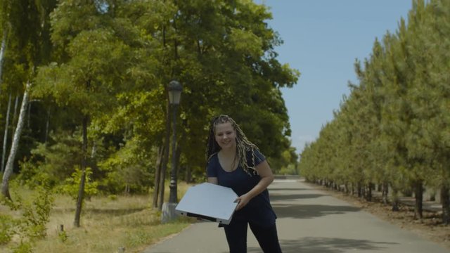 Active young woman rollerblading at speed with pizza box working in fast pizza delivery service in public park. Positive female riding rollerblades along park alley hurry deliver pizza to customer.