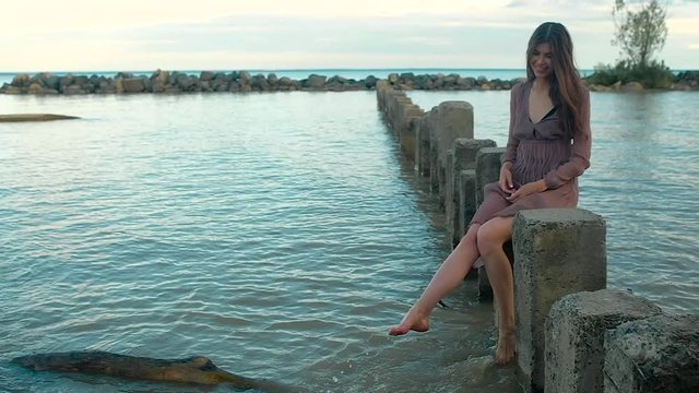 young girl in transparent brown dress, barefoot gracefully sits on stone rectangular pillars standing in sea, dangles her legs making splashes. straightens his long black hair, smiles coquettishly.