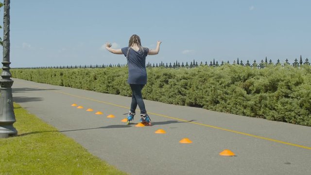 Rear view of skillful woman rollerblading inline roller skates past cones on park footpath. Active female with ponytail from afro-braids training skill of crisscross ride during inline slalom outdoor.