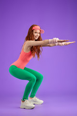 Joyful young red-haired woman in comfortable aerobics clothes of 80s style models and poses in...