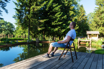 Attractive young man in glasses, t-shirt and shorts greets the dawn while sitting in front of a pond on a wooden chair