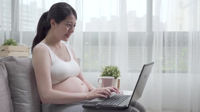 Young beautiful pregnant woman freelance working on laptop with tea at home office. future mom worker replying customer on internet typing notebook computer during startup small business online shop.