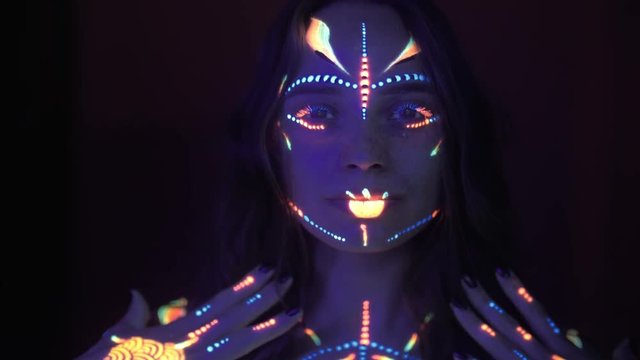 Portrait of woman with ethnic pattern, neon makeup in ultraviolet light. Fluorescent paint and pencil are on the table. Body Art design of female posing in UV, painted face, colourful make up, over da
