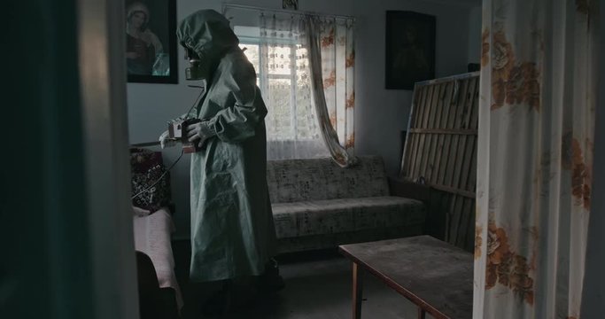 Unrecognizable male in hazmat suit and gas mask using Geiger counter on old furniture inside abandoned house in Chernobyl.