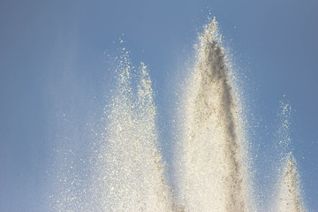 Slightly defocused jets and splashes of water rising sharply and high. Working refreshing fountain in a public park in the city. Aqua show. Splatter of water against the blue clear sky.